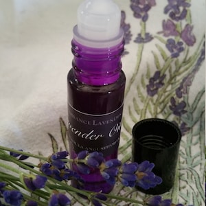 May include: A purple glass roller bottle with a white roller ball. The bottle is labeled "Essence Lavender Oil Lavandula Angustifolia". The bottle is sitting on a white fabric with purple lavender flowers.