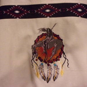 May include: Embroidered design of a Native American warrior riding a horse, holding a spear, with a red, orange, and yellow background. The design is surrounded by a dark red circle with a black border. Five white feathers with yellow accents hang from the bottom of the circle.