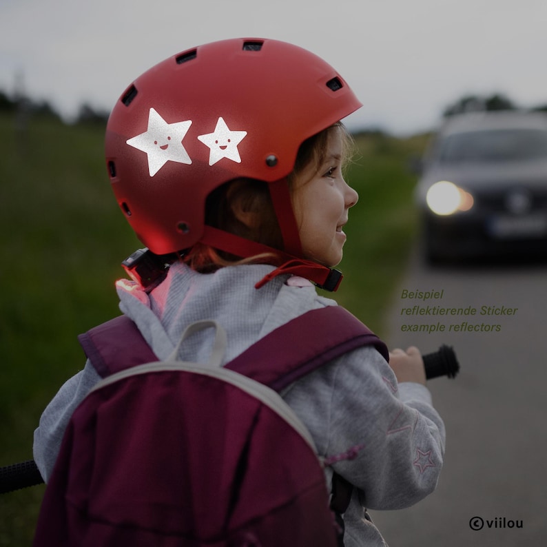 Pu&ograve; includere: Un casco da bicicletta rosso con due adesivi riflettenti bianchi a forma di stella con faccine sorridenti sul lato. Il casco presenta diverse fessure di ventilazione e un cinturino sottogola rosso. Il testo "Beispiel reflektierende Sticker example reflectors" &egrave; visibile.