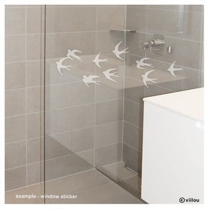 Pu&ograve; includere: Un set di adesivi bianchi a forma di rondine applicati su una parete doccia in vetro. Il design presenta otto sagome di uccelli in diverse posizioni di volo, creando un effetto stormo. Il testo "example - window sticker" e "viilou" sono visibili.