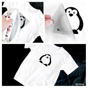 Puede incluir: Una camiseta blanca con un dise&ntilde;o de ping&uuml;ino negro planchado. El ping&uuml;ino est&aacute; mirando hacia adelante con las alas extendidas y tiene un peque&ntilde;o pico y ojos.