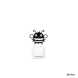 May include: A black and white cartoon bee sticker sits above a white light switch. The bee features a smiling face, antennae, and a striped body. The image is set against a plain white background.