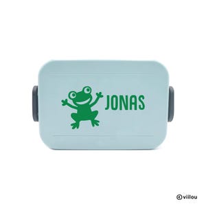 Puede incluir: Una fiambrera azul claro con un dibujo de rana verde y el nombre "JONAS" impreso en verde. La fiambrera tiene cierres laterales negros y una forma rectangular redondeada. La rana sonríe con los brazos extendidos.