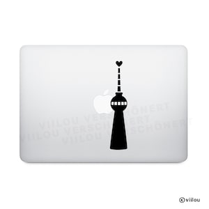 May include: A black vinyl sticker featuring the silhouette of the Berlin TV Tower with a heart above it. The sticker is placed on a white laptop, with the Apple logo visible. The design is simple and modern.
