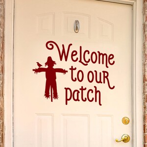 May include: A white door with a burgundy decal that reads "Welcome to our patch." The decal features a scarecrow with a bird perched on its arm. The door has a gold-colored handle and is surrounded by a brick facade.