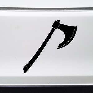 May include: Black axe decal, a vinyl sticker with a stylized axe design. The axe features a dark blade and a long handle, suitable for application on vehicles or other surfaces. The decal is a simple, bold graphic.