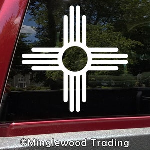 May include: White vinyl decal of the Zia symbol, a traditional symbol of the Pueblo people of New Mexico, on a red car window. The symbol is a stylized sun with four rays, each with four lines.