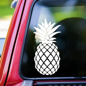 May include: White vinyl decal of a pineapple with a detailed design. The decal is on a red car window.