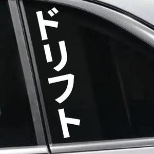 May include: White vinyl decal with Japanese characters for "drift" applied to a car window. The decal is vertically oriented and placed on the rear passenger window of a silver car. The text is in a bold, sans-serif font.