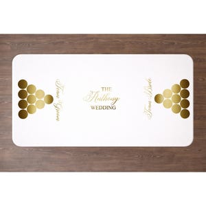 May include: A white rectangular table cover with gold accents. The cover features gold beer pong ball arrangements on each end, with the words "Team Groom" and "Team Bride" in gold script. The center displays "The Anthony Wedding" in elegant gold script.