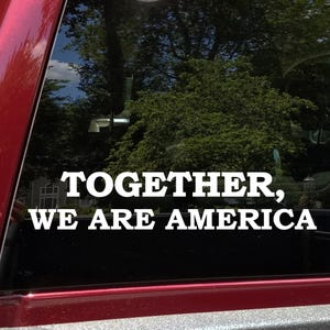 May include: White decal with the text "TOGETHER, WE ARE AMERICA" on a vehicle window. The background shows a lush green tree line and a glimpse of a house. The decal is placed on a red surface.