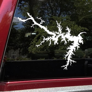 May include: White decal of a lake outline, applied to a vehicle window. The decal's design mimics a detailed map of the lake's shoreline, with intricate branches and inlets. The background shows trees and a glimpse of the sky.