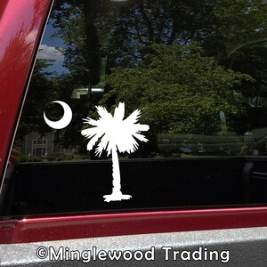 May include: White vinyl decal of the South Carolina state flag, featuring a palmetto tree and a crescent moon. The decal is displayed on a red truck window.