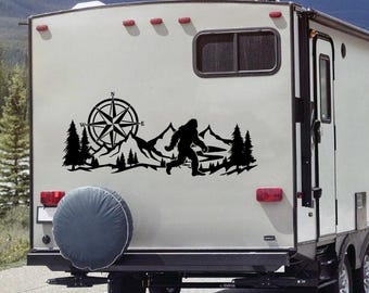 May include: Black vinyl decal featuring a compass rose, mountains, pine trees, and a silhouette of Bigfoot running. The decal is applied to the back of a recreational vehicle, creating a nature-themed design.