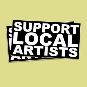 May include: Two black rectangular stickers with white text that reads "SUPPORT LOCAL ARTISTS" are layered on a light yellow background. The stickers have a white border.