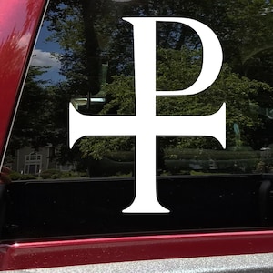 May include: A white Chi Rho symbol decal, a combination of the first two letters of the Greek word for Christ, is affixed to a vehicle window. The symbol is a cross with the letter 'P' superimposed on it. The background shows trees and a house.