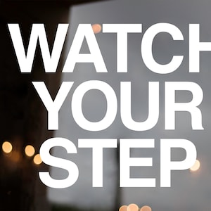 May include: A white sign with black text that reads "WATCH YOUR STEP." The sign is on a glass door with a wooden frame. The background is blurry and out of focus.