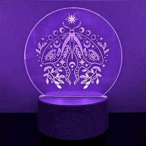 May include: A round, clear acrylic night light with a detailed moth design, illuminated by a purple LED. The moth illustration includes crescent moons, stars, and floral elements. The light sits on a natural wood base, creating a mystical ambiance.