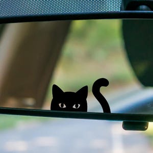 May include: A black cat peeking over a car's rearview mirror. The cat's head and tail are visible, with its eyes and ears clearly defined. The sticker is a simple, minimalist design.