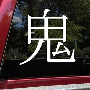 May include: White vinyl decal featuring the Japanese kanji character for "demon" or "oni." The character is displayed in a bold, stylized font. The decal is affixed to a window, with a red vehicle frame visible.