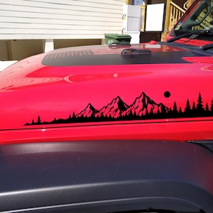 May include: A black vinyl decal applied to the red hood of a vehicle. The decal features a detailed silhouette of a mountain range and a dense forest of pine trees, with a small circular sun positioned above the peaks on the right side.