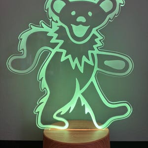 May include: A green glowing acrylic bear silhouette on a wooden base. The bear is walking with its arms outstretched and has a smiling face.