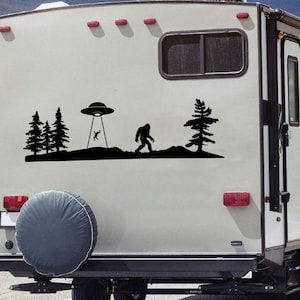 May include: A white camper van with a black silhouette decal of a UFO, Bigfoot, and trees. The decal is on the back of the van.