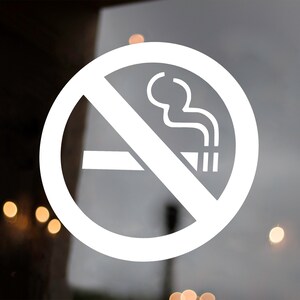 May include: A white, circular no smoking sign with a cigarette and smoke symbol, crossed out by a diagonal line. The sign is on a glass door, with a blurred background and wooden door frame.