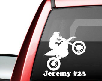 May include: White silhouette of a person riding a dirt bike with the name Jeremy #23 printed below. The decal is on the back window of a red pickup truck.