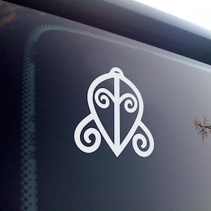 May include: White vinyl decal featuring a stylized, symmetrical design. The graphic is a teardrop shape with a central vertical line and swirling details. The decal is affixed to a dark, reflective surface, likely a car window.