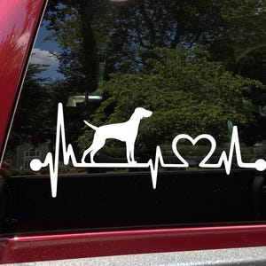 May include: White vinyl decal of a dog silhouette with a heartbeat line and a heart. The decal is on a car window.