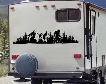 May include: Black vinyl decal featuring a silhouette of a mountain range, forest, and Bigfoot walking. The decal is applied to the back of a recreational vehicle, with a spare tire cover visible below. The design is a solid black color.