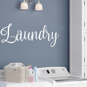 May include: A white laundry room sign with the word "Laundry" in cursive script. A white washing machine with a glass lid is in the foreground. A wicker laundry basket with folded clothes is on the counter next to two bottles of laundry detergent.