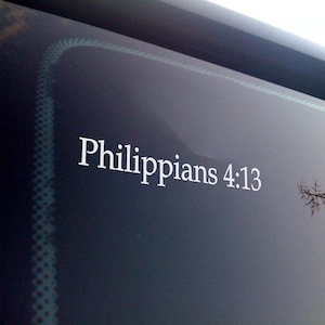 May include: White text on a dark background reads "Philippians 4:13". The text is in a serif font. The background appears to be a car windshield.