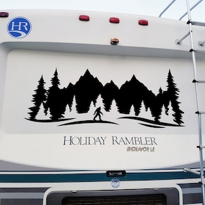 May include: A white recreational vehicle with a black silhouette decal of a forest scene with mountains, trees, and a Bigfoot figure. The decal is on the back of the vehicle, which also has the text "HOLIDAY RAMBLER ENDEAVOR LE" in black lettering.