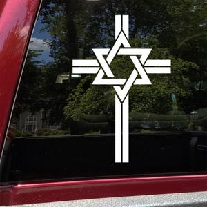 May include: White vinyl decal featuring a cross with a Star of David at the center. The decal is applied to a vehicle window, with a red frame visible. The background shows trees and a house.