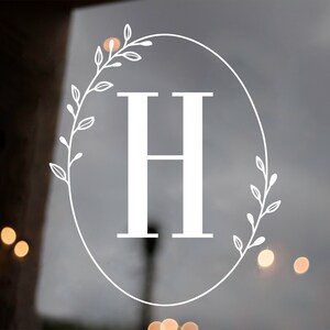 May include: White vinyl decal with a wreath of leaves and the letter H in the center. The decal is on a glass surface.