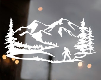 May include: White vinyl decal of a mountain range with a silhouette of a Bigfoot walking in the foreground. The mountains are in the background with a river flowing through the valley. There are trees on either side of the river.