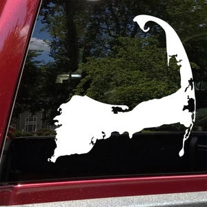 May include: A white vinyl decal of the Cape Cod peninsula, affixed to a vehicle window. The decal's shape is a simplified outline of the landmass, with detailed cutouts along the coastline. The background shows trees and a glimpse of a building.