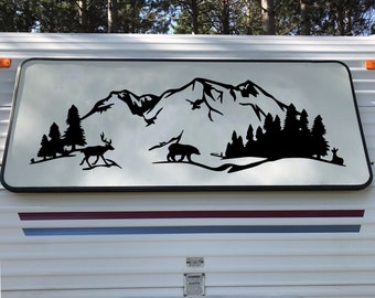 May include: Black and white silhouette decal of a mountain range with trees, a bear, and a deer. The decal is on a white camper trailer.