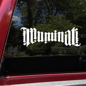 May include: White vinyl decal with the word "Illuminati" in a gothic font, affixed to a vehicle window. The decal is visible against a backdrop of trees and a glimpse of a house.