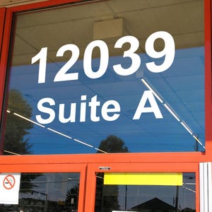 May include: Exterior shot of a storefront with large white numbers "12039" and the words "Suite A" on the glass. The door frame is orange, and the sky is blue. A no-smoking sign is visible.