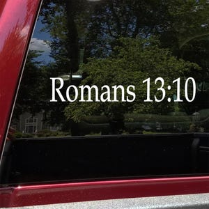 May include: White text on a vehicle window reads "Romans 13:10". The text is centered and appears to be a decal or sticker. The background shows trees and a glimpse of a house, with the vehicle's red paint visible on the left.