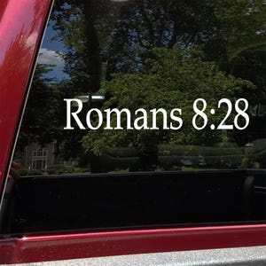 May include: White decal with the text "Romans 8:28" on a vehicle window. The text is in a clean, sans-serif font. The background shows a blurred view of trees and a house.