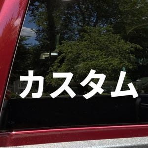 May include: White vinyl decal with Japanese characters, "カスタム", on a car window. The decal is in a bold, sans-serif font. The background shows a blurred view of trees and a building.