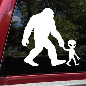 May include: White vinyl decal featuring a Bigfoot figure holding hands with an alien. The figures are walking together, with the Bigfoot in the lead. The decal is designed to be applied to a vehicle window.