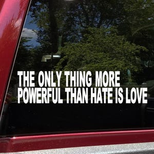 May include: White vinyl decal with the text "THE ONLY THING MORE POWERFUL THAN HATE IS LOVE" in a bold, sans-serif font. The decal is displayed on a window with a blurred background of trees and sky.