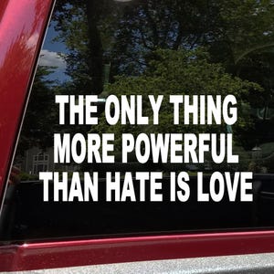 May include: White vinyl decal with the text "THE ONLY THING MORE POWERFUL THAN HATE IS LOVE" on a car window. The decal is in a bold, sans-serif font. The background shows trees and a house.