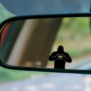 May include: A black Bigfoot silhouette with a heart-shaped hand gesture is reflected in a car's rearview mirror. The image is set against a blurred background of the car's interior and exterior.