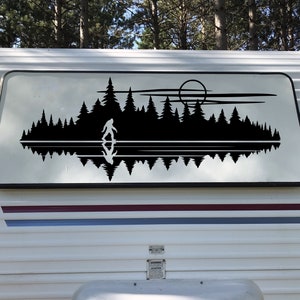 May include: A black vinyl decal featuring a silhouette of Bigfoot standing in front of a forest and its reflection in the water, with a sun and clouds in the background. The decal is applied to a white RV panel.